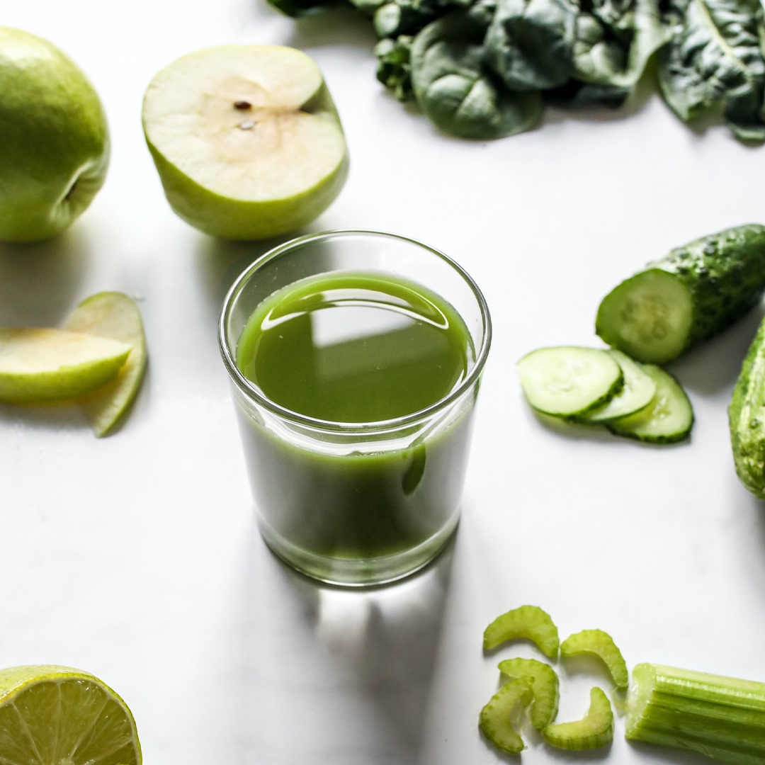 A glass of green juice surrounded by green apple, cucumber slices, celery, leafy greens and lime.
