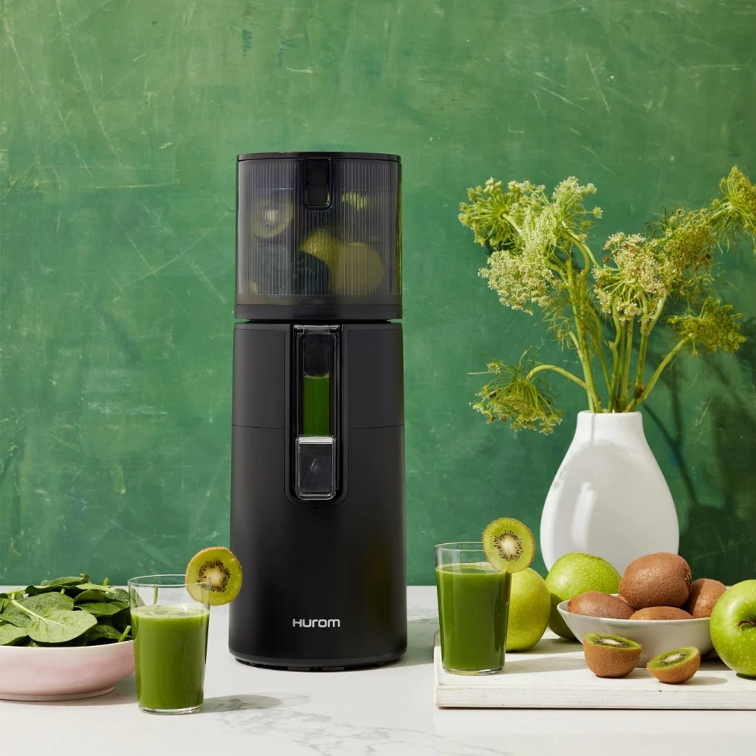 A black Hurom juicer beside two glasses of green juice that are garnished with kiwi slices, with leafy greens, apples and kiwis on the worktop.