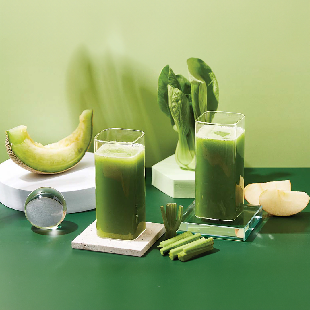 Two tall glasses of green juice beside a melon wedge, apple slices, celery sticks and leafy greens on a green worktop.