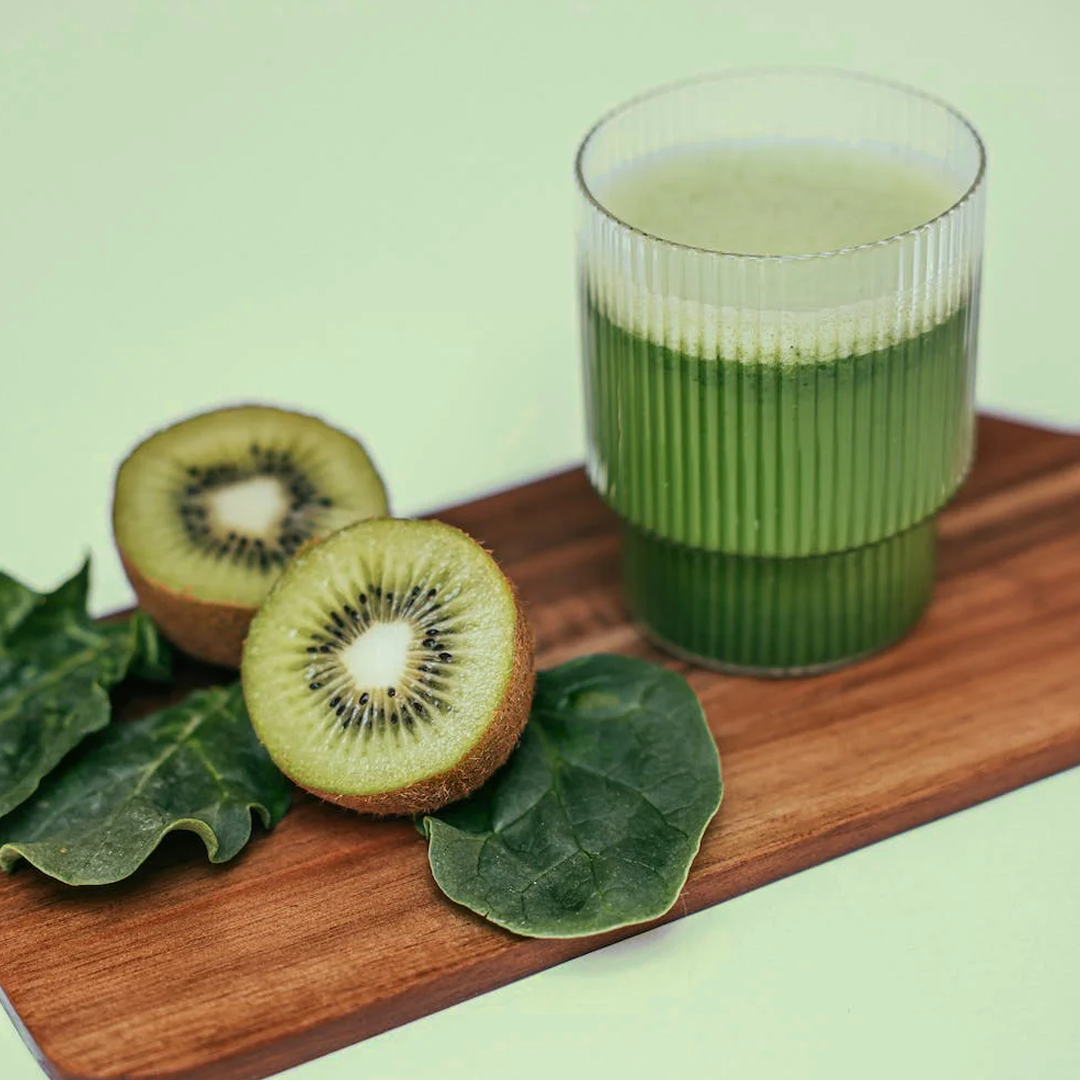 A glass of frothy green juice beside kiwi halves and spinach leaves on a wooden board.